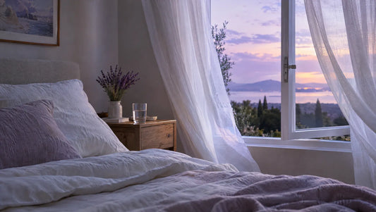 A cool, airy bedroom in evening light with an open window, sheer curtains and a glass of water by the bed
